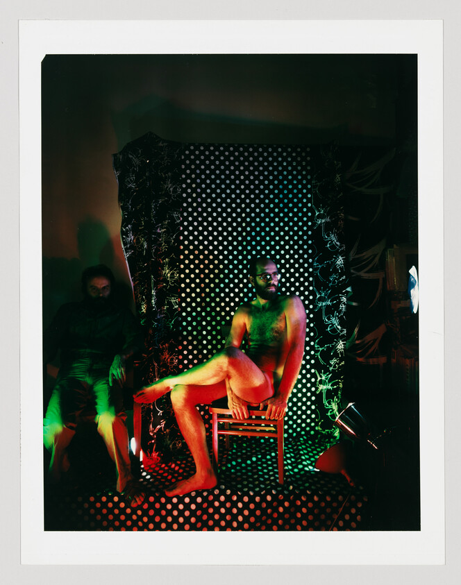 Bearded man sits nude on a chair with crossed legs under red and green stage lighting.