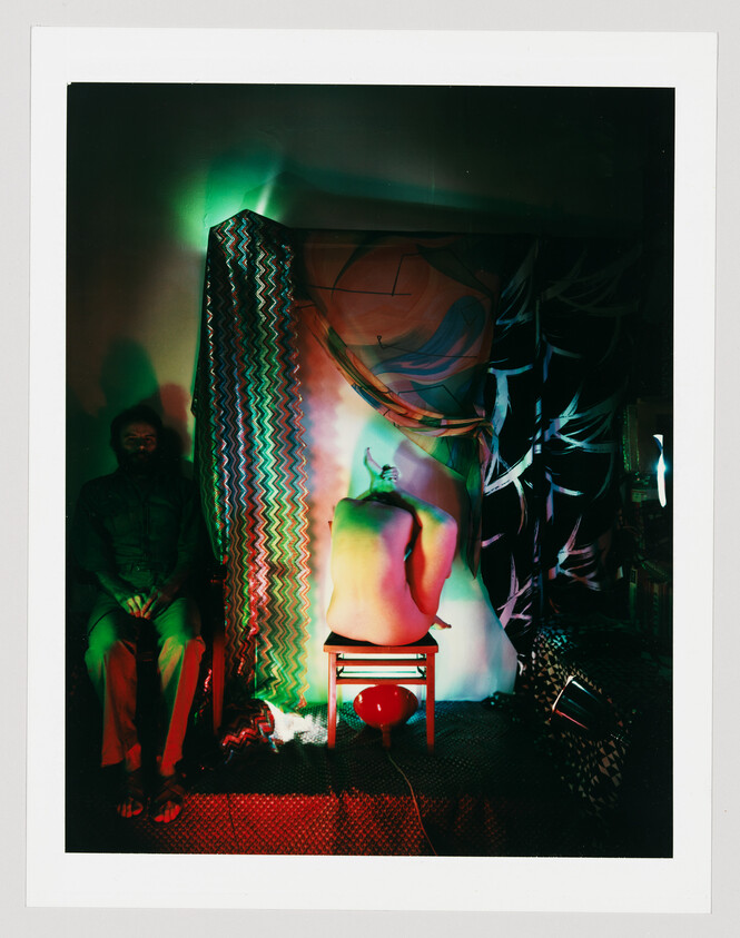 A naked person sits on a small stool with their back turned under colorful stage lighting.