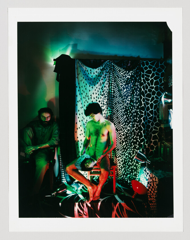 A shirtless man sits on a chair holding a mask and looking down under green and red lights.