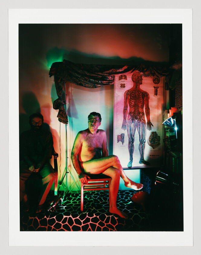 A shirtless man sits cross-legged on a chair under colored lights beside a large anatomical chart.