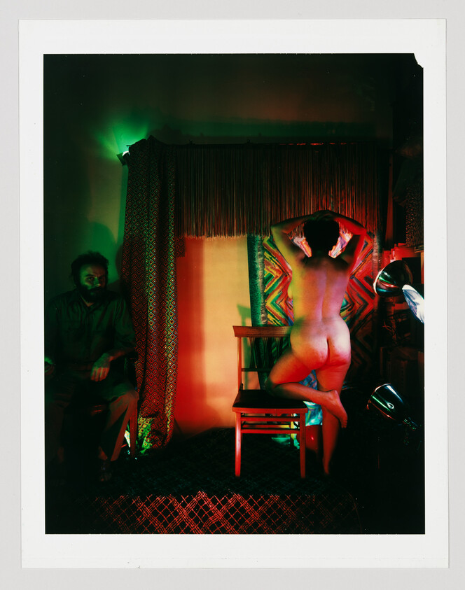 A nude person stands on a chair posing with hands behind their head while another sits in shadow.