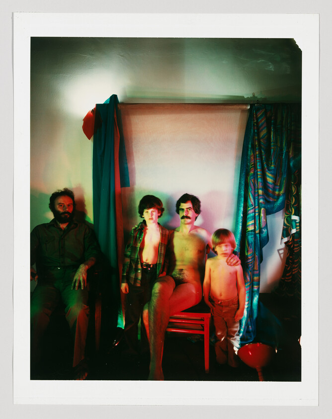 Four people pose against a draped backdrop, a seated mustached man with two shirtless boys and a bearded man.