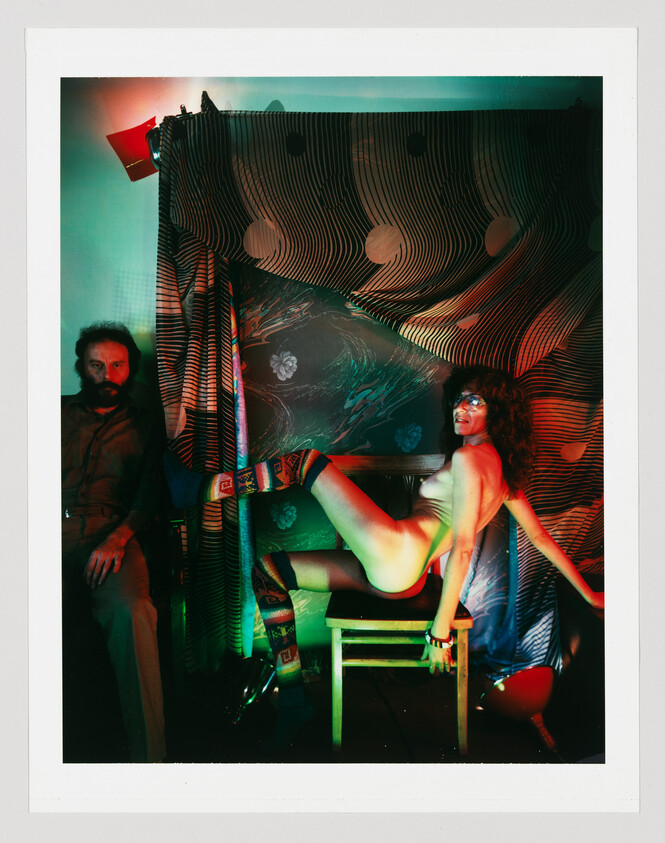 Woman wearing colorful knee socks poses nude on a chair with a seated man nearby.