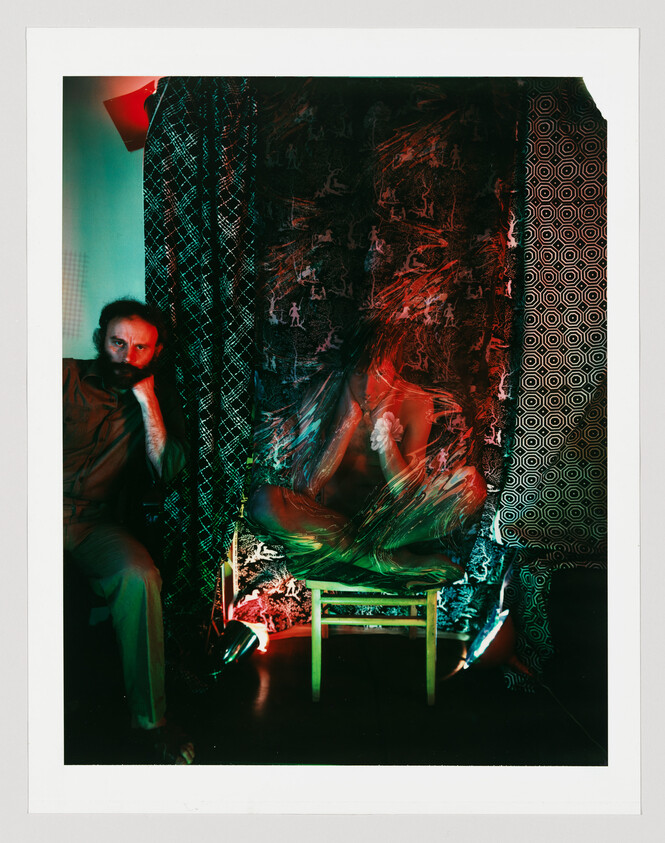 A bearded man watches a blurred person sitting cross-legged on a brightly lit chair.