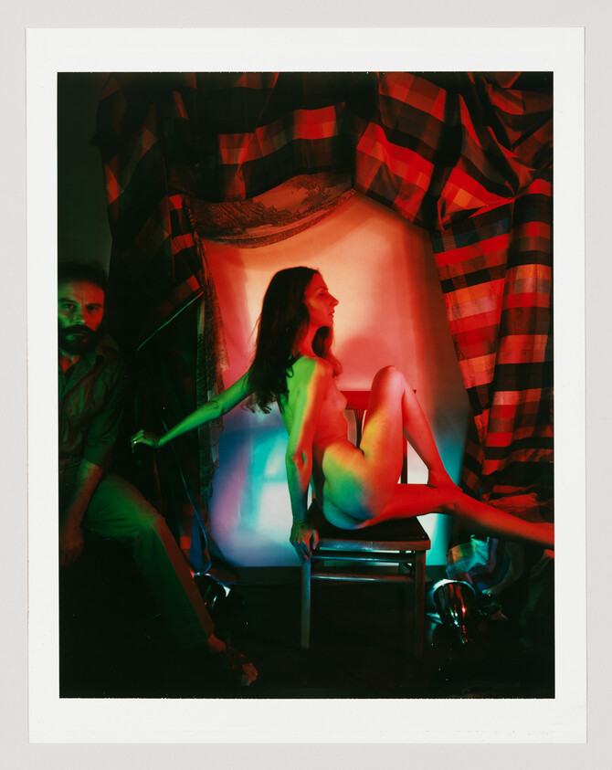 Nude woman posed on a chair under red and green theatrical lighting while a man stands nearby.