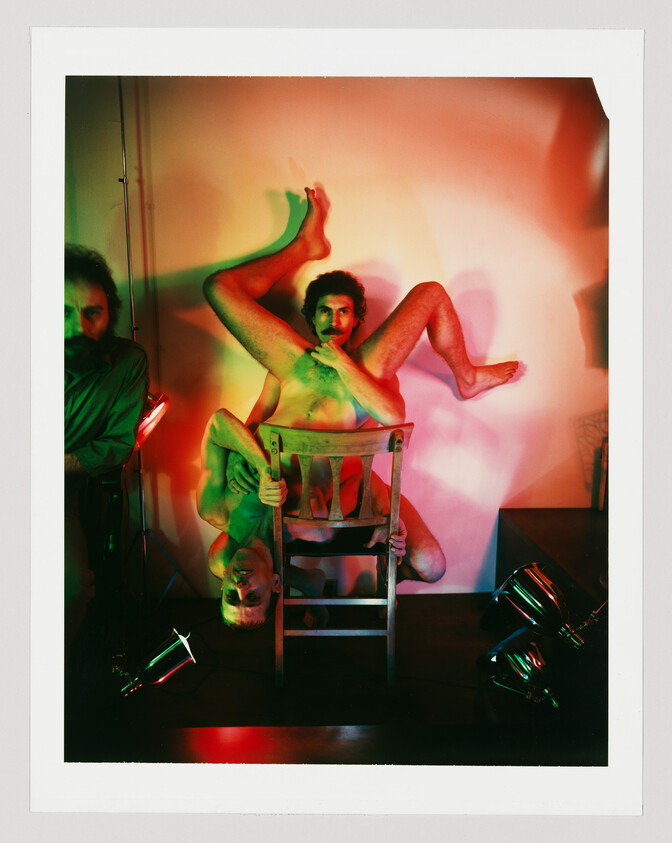 Two men pose naked around a wooden chair, one upside down with legs raised against a colored background.