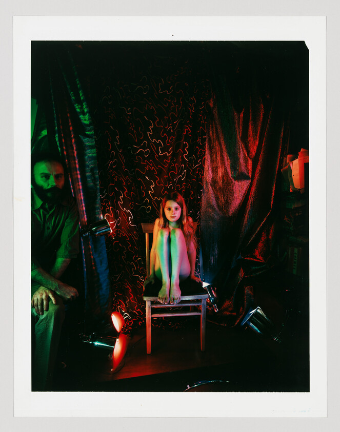 A young girl sits on a chair with her knees pulled to her chest under colored studio lights.