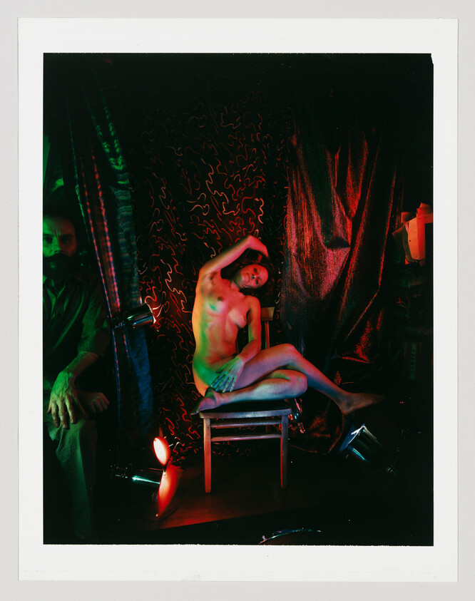 A nude model poses gracefully on a chair under colorful stage lighting while a man watches.