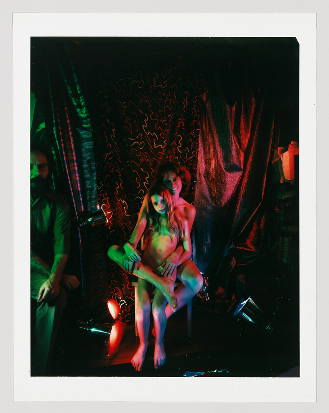 Two people sit closely with one person seated on the other's lap under colored stage lights.