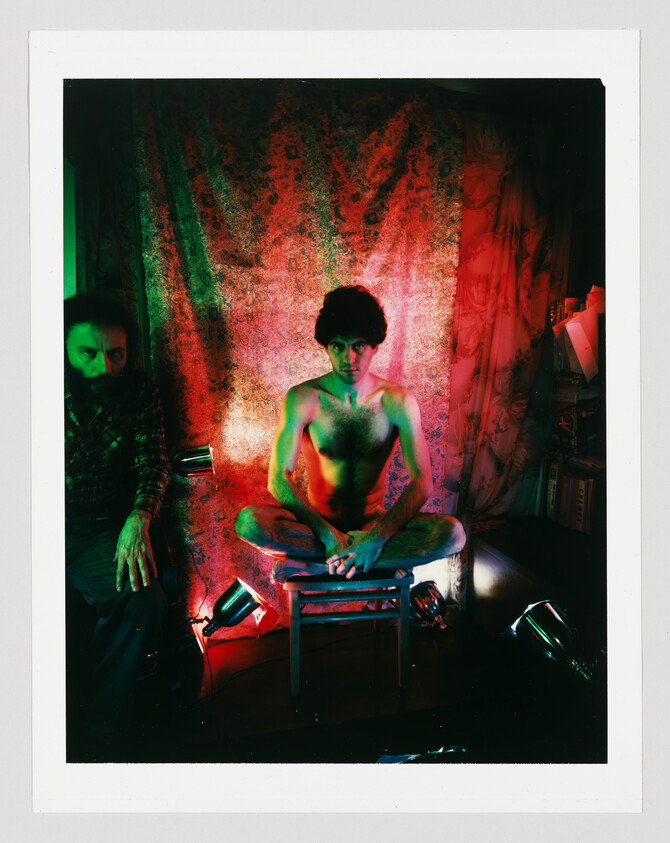 Shirtless man sits cross-legged on a small stool under red and green lights while another man watches.