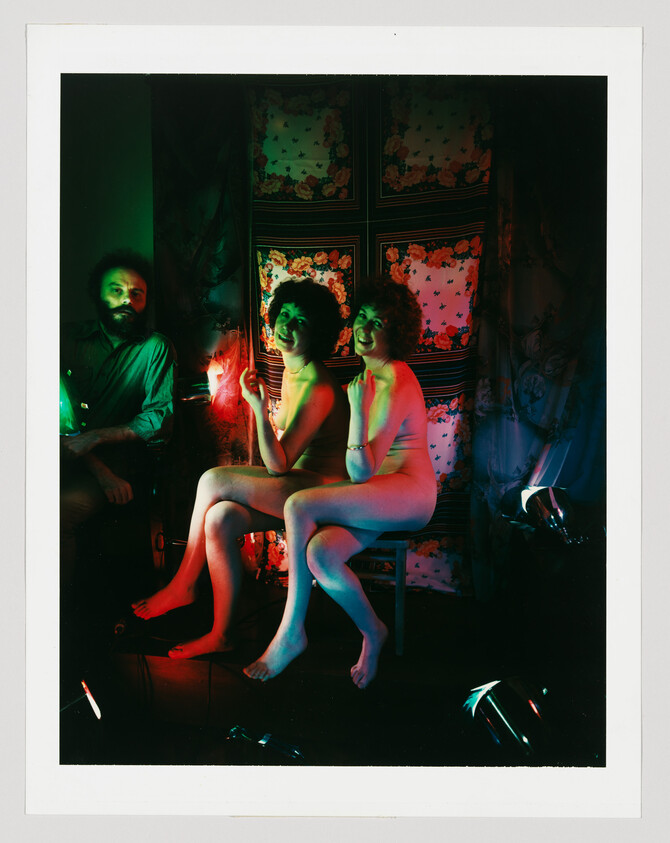 Two nude women sit together on a stool smiling under red and green lights while a man watches.