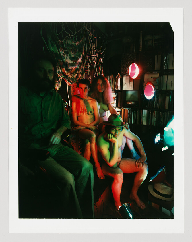 Three mostly nude people and one clothed person sit together in a dim room lit by colored lights.