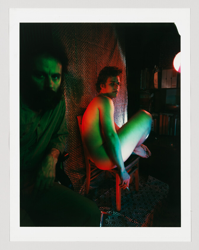 A naked man crouches on a chair under red and green lighting while a bearded man watches.