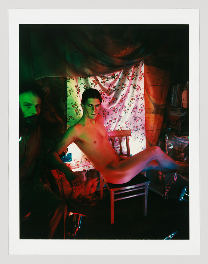 Young man posing nude draped across a wooden chair in a dim room with floral curtain.