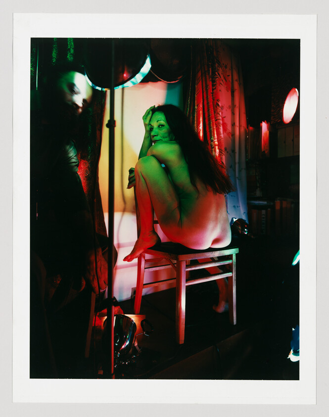 A nude woman crouches on a stool under colorful red and green stage lighting, looking toward the camera.