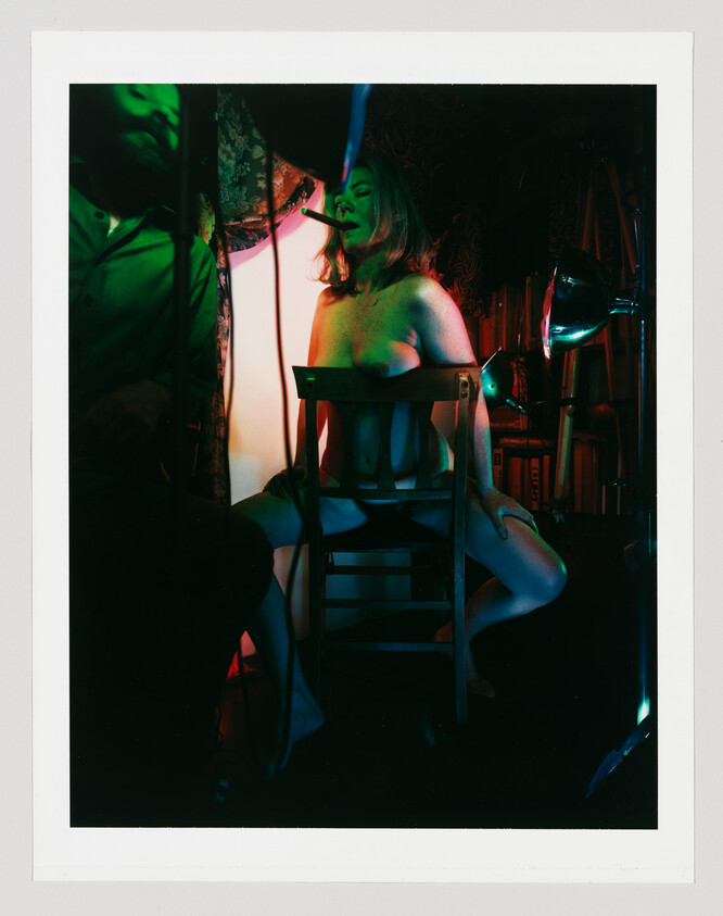 A topless woman sits on a wooden chair with legs spread, holding a cigarette in her mouth.