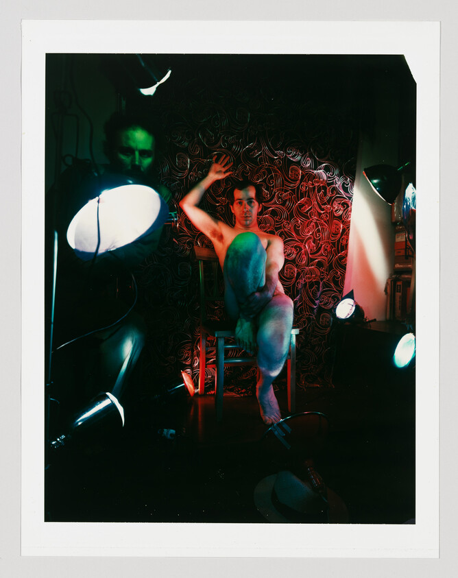Nude man sits on a chair with one knee raised, posing under colored studio lights.