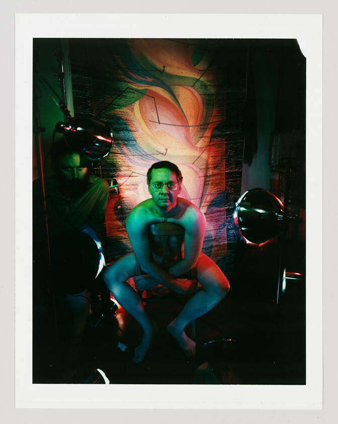 A naked man sits on a chair under green and red lights in front of a painted backdrop.