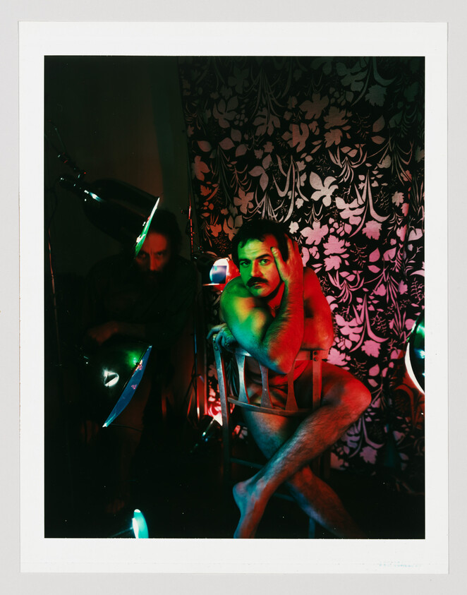 A shirtless man with a mustache poses on a chair under green and red lights with a floral backdrop.