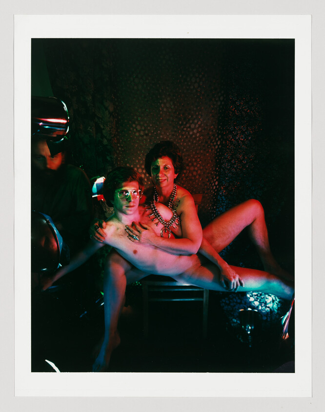 Woman wearing jewelry holds a nearly naked young man reclining across her lap under colored lights.