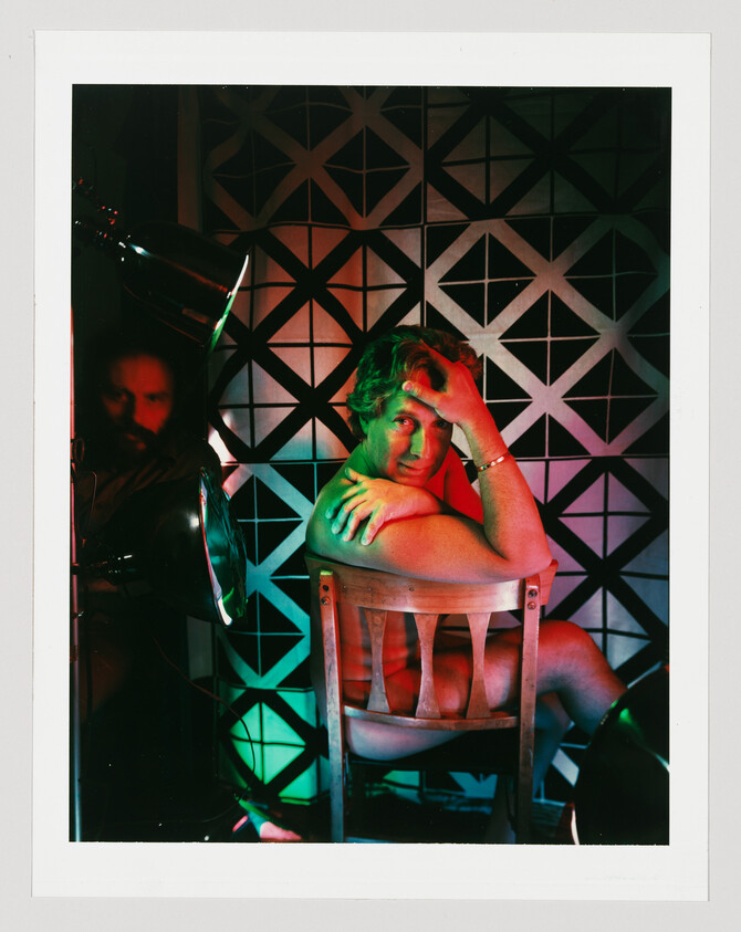 A shirtless man sits backward on a wooden chair under green and red studio lights, hand on forehead.