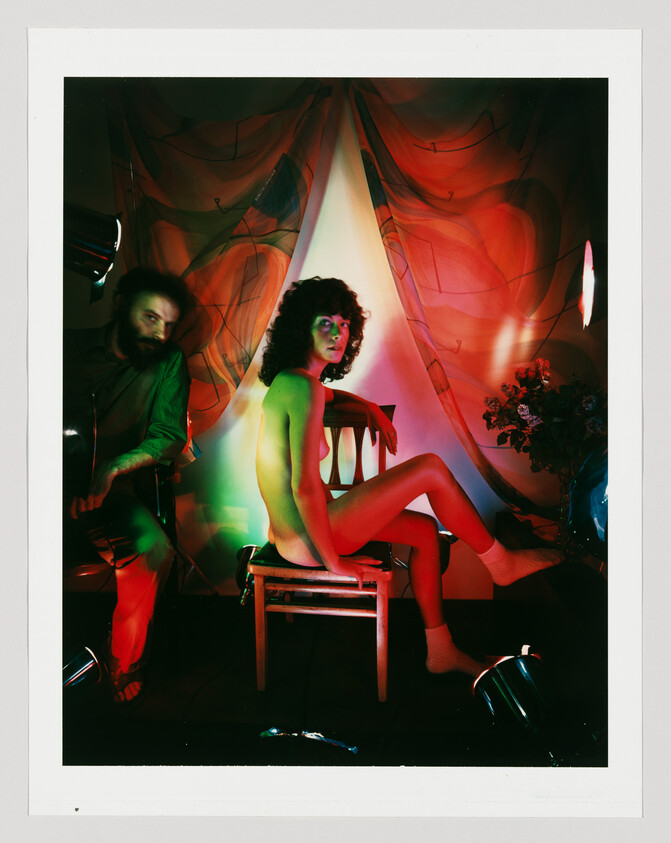 A naked woman sits on a wooden chair under colorful stage lights while a man adjusts lighting.