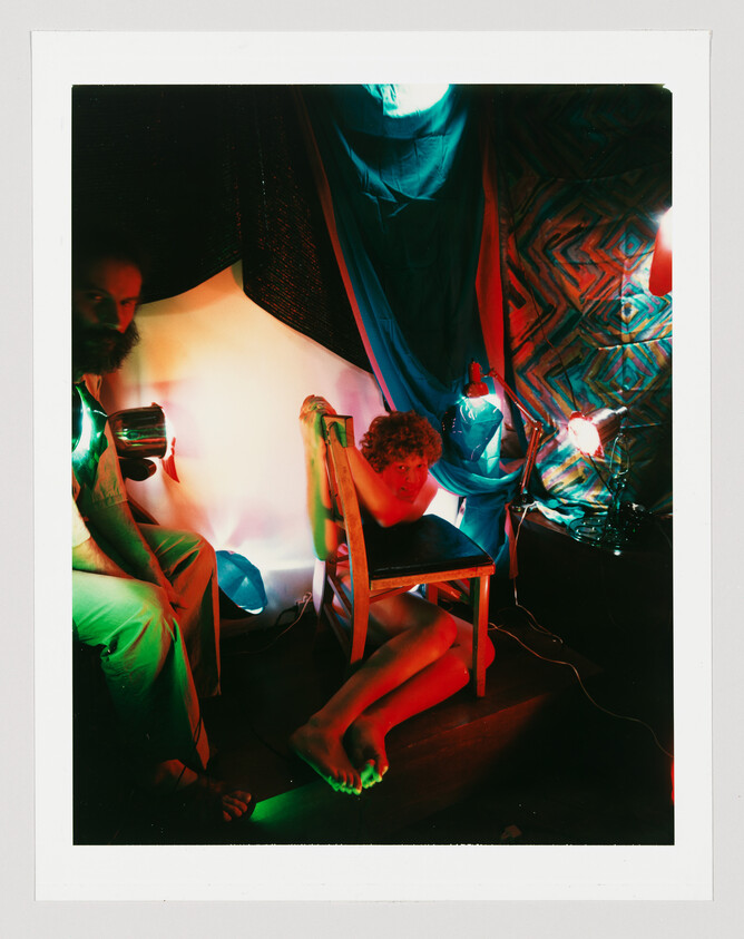 A young person crouches behind a wooden chair under colorful red and green lights while a bearded man watches.