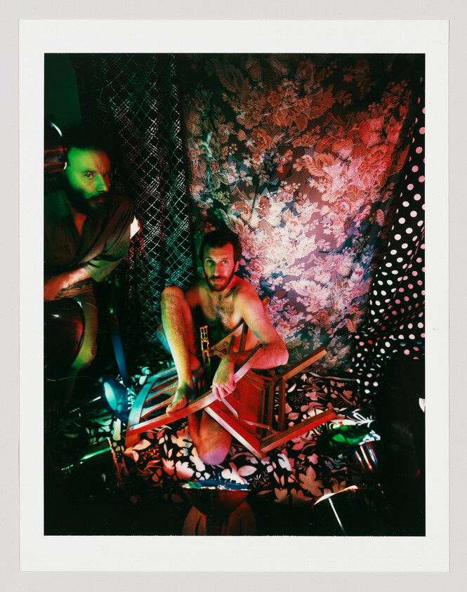 Shirtless man sits amid patterned fabrics holding broken chair pieces while another man watches.