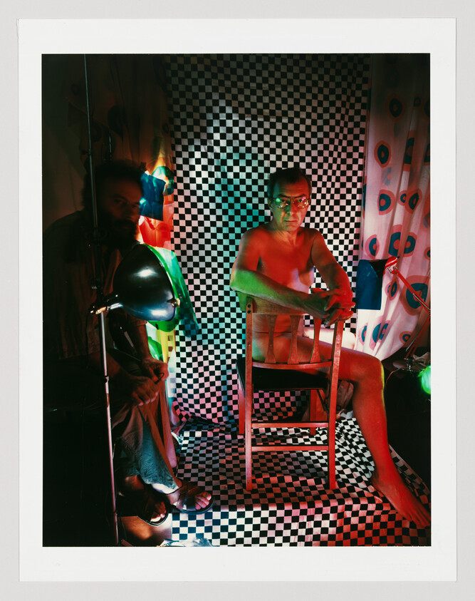 A shirtless man sits on a wooden chair against a black-and-white checker backdrop lit by colorful lamps.
