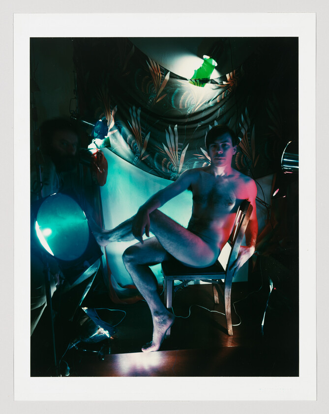 A nude man sits cross-legged on a chair amid colored studio lights and a patterned backdrop.