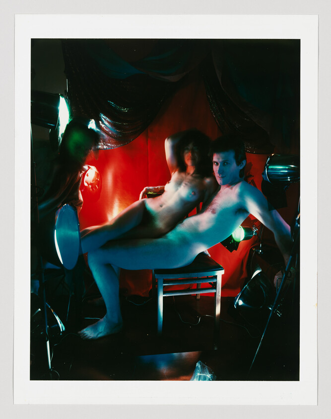 Two nude people pose dramatically on a chair amid colored studio lights and red backdrop.