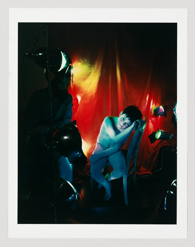 A young person sits nude on a chair, leaning their head on their hand amid colored studio lights.