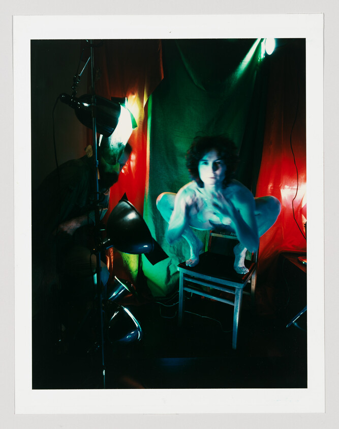 Person crouching on a chair under colored studio lights, hands raised toward the camera.