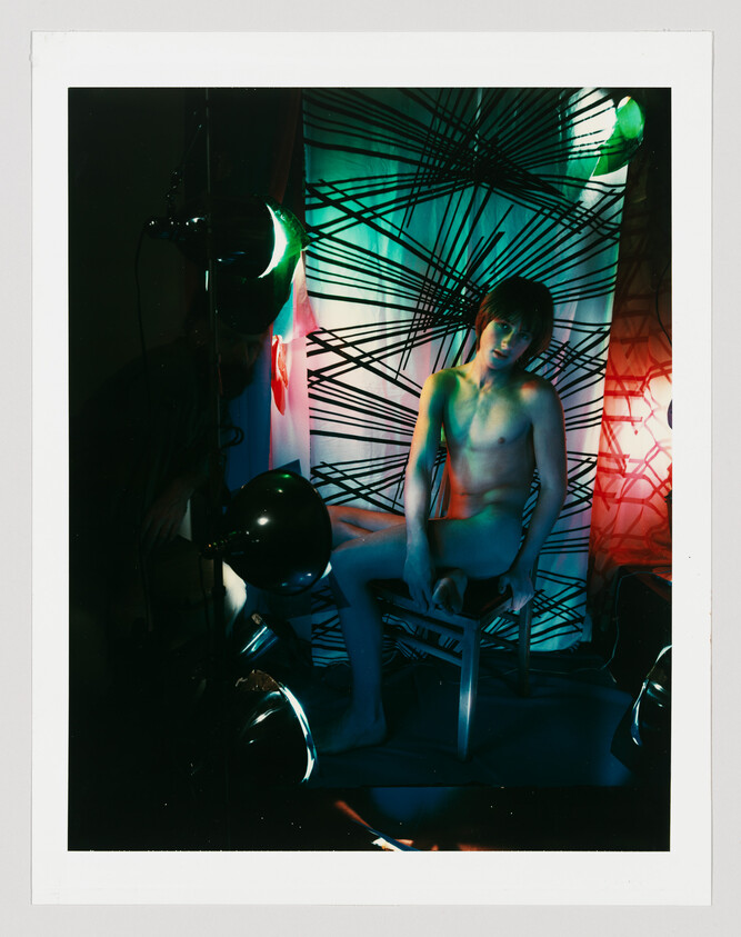 Nude person sitting on a stool surrounded by colored lamps and a black-striped backdrop.