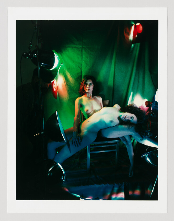 Two nude women posed among studio lights and green backdrop, one reclining across the other's lap.