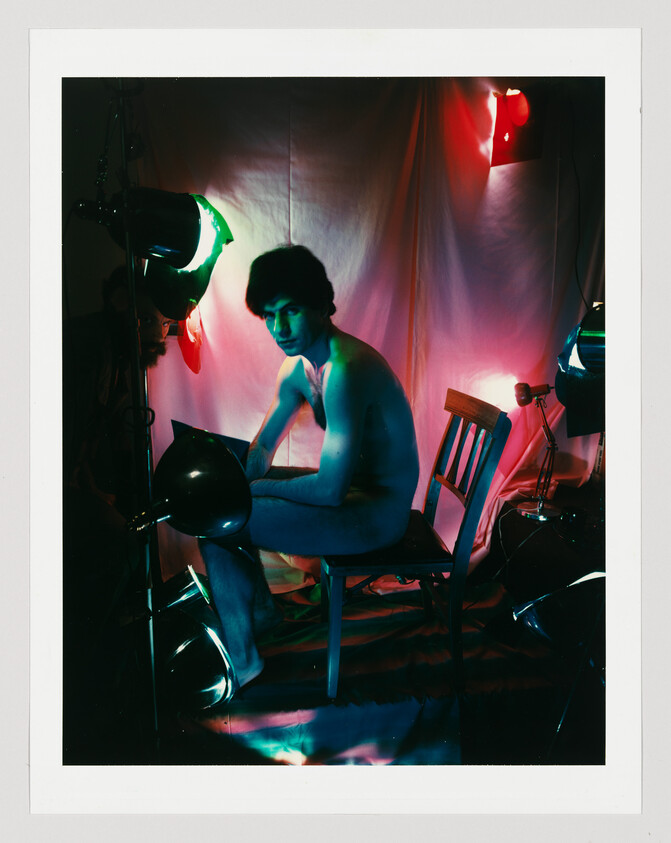 Naked person sitting on a wooden chair in a studio surrounded by colorful spotlights.