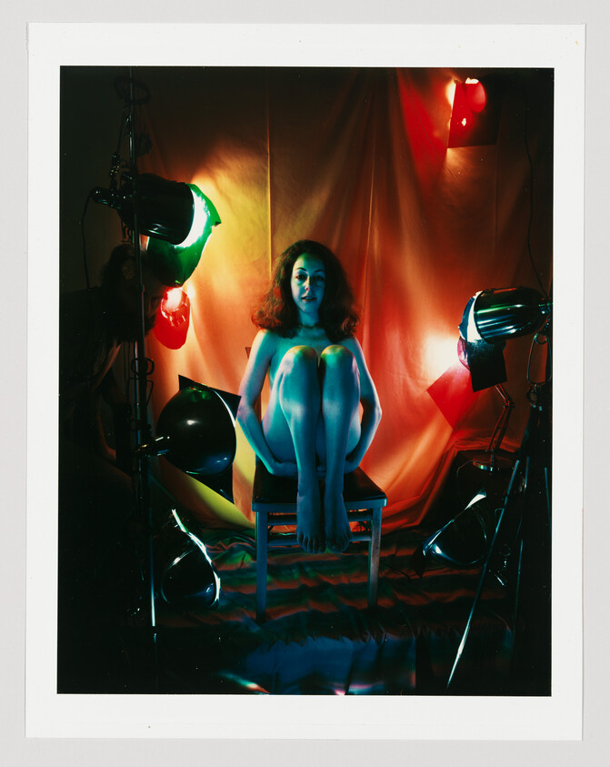 A woman sits barefoot on a chair hugging her knees under colorful studio lights.