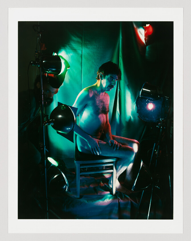 A naked man sits on a chair surrounded by colored studio lights and draped fabric.