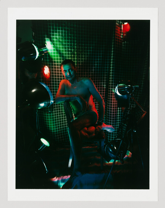 A shirtless man sits on a chair under green and red studio lights against a grid backdrop.