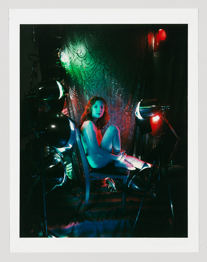 A woman sits curled on a chair surrounded by green and red studio lights and lamps.