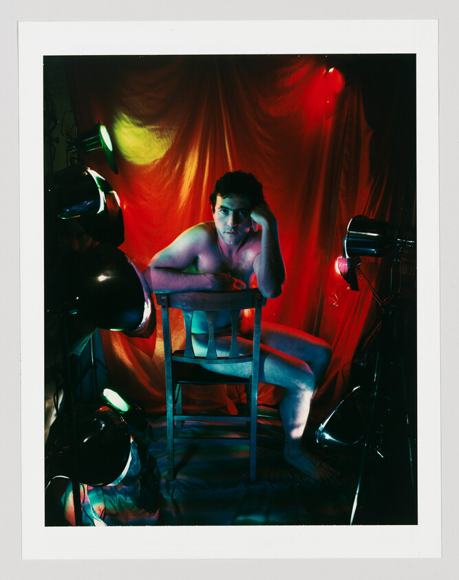 A nude man sitting backwards on a wooden chair under colorful studio lights.