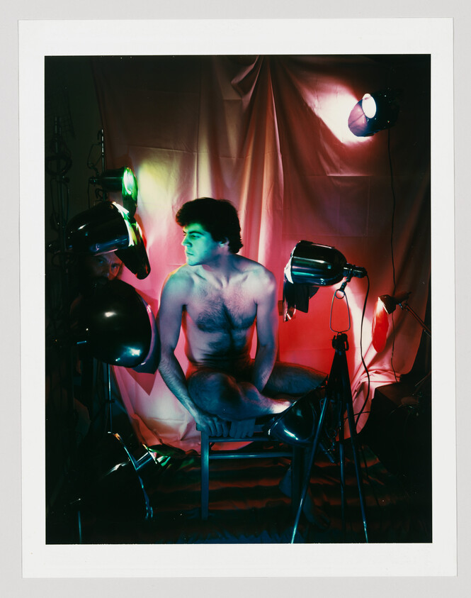 A shirtless man sits cross-legged on a stool surrounded by colorful studio lights and draped backdrop.