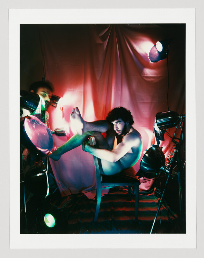 A shirtless man sits on a chair with his legs raised, surrounded by colorful studio lights.