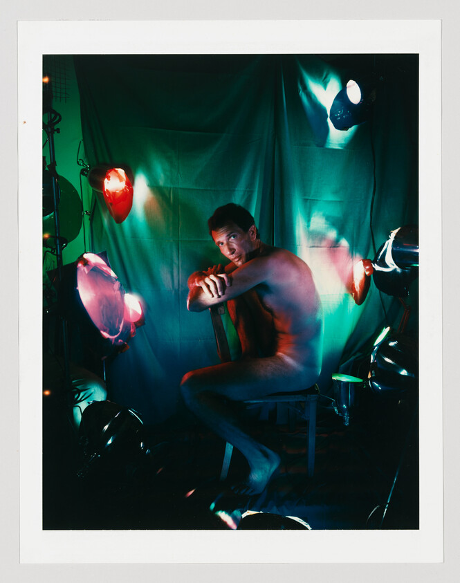 A nude man sits on a stool amid colored studio lights and a green backdrop.