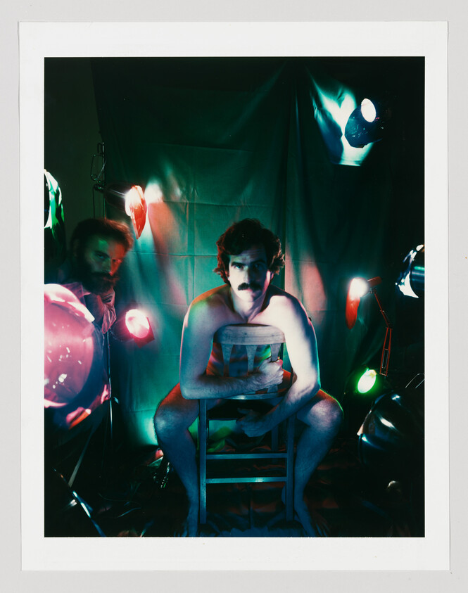Man with mustache sitting barefoot backward on a chair under colorful studio lights, another man peeking.