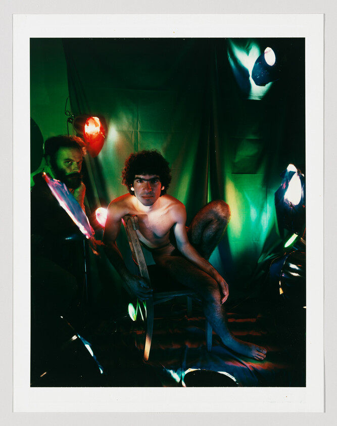 A shirtless man crouches on a wooden chair under dramatic green and red studio lights while an assistant adjusts lighting.