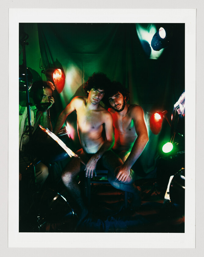 Two shirtless men sit close together under red and green stage lights while a person adjusts lighting.