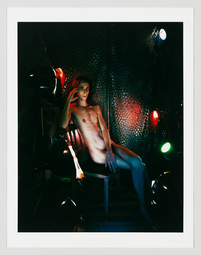Young person seated nude on chair, posing under colored stage lights against a dark patterned backdrop.