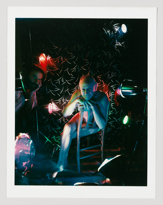 An older man sits on a wooden chair under colorful stage lights, leaning forward and resting on a cane.
