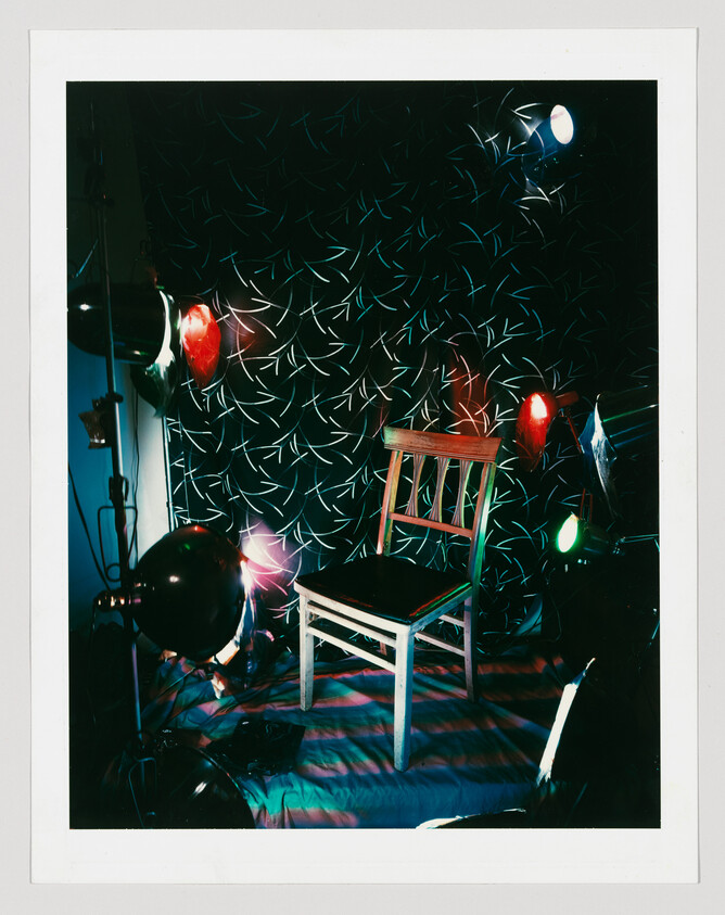 A wooden chair centered in a dark room lit by colorful studio lights and patterned wallpaper.
